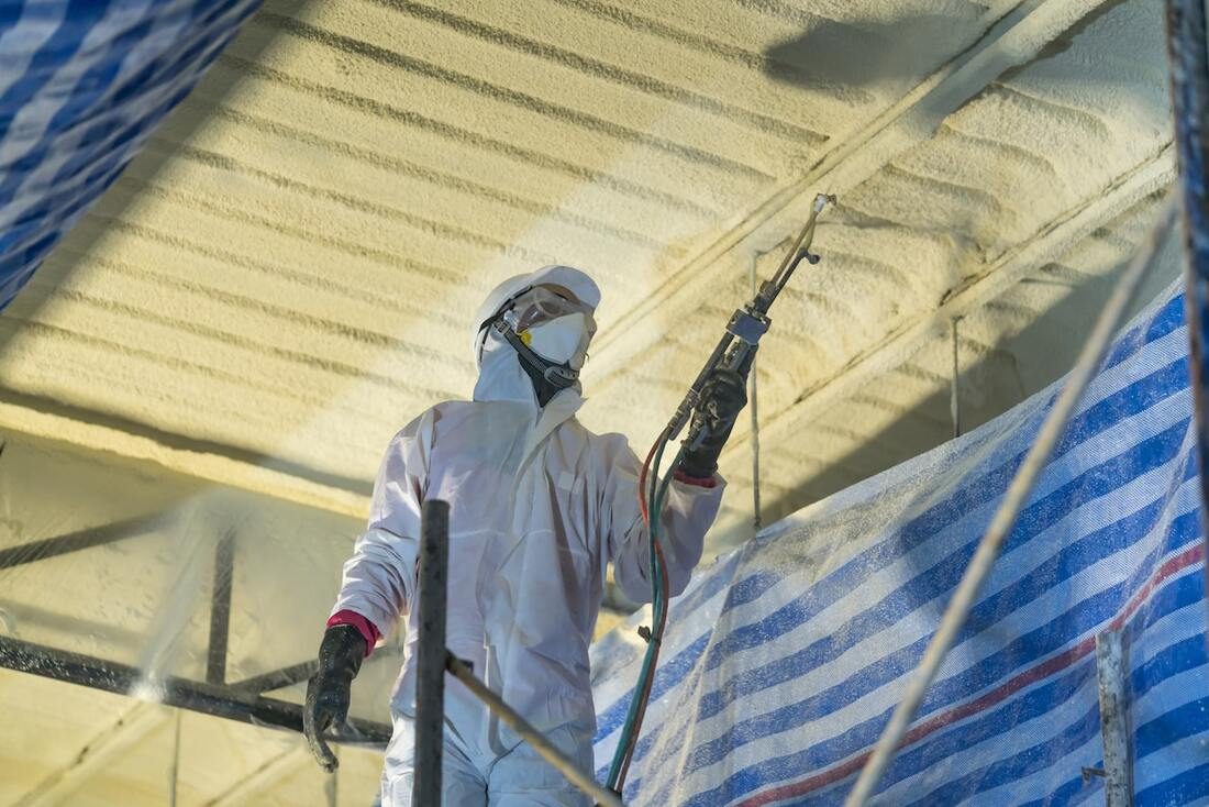 Spray foam technician in protective gear applying closed-cell foam to a ceiling