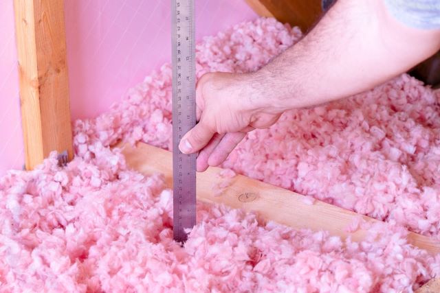 Installer measuring the depth of pink blown-in insulation between attic joists with a ruler